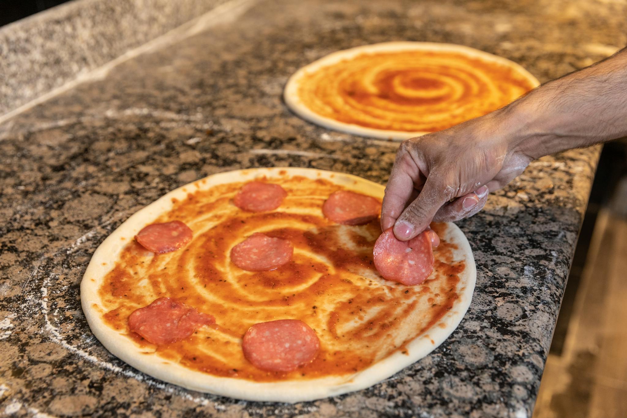 A hand placing pepperoni slices on pizza dough with tomato sauce.